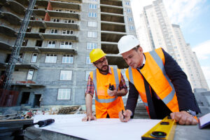 Engenheiro moderno apontando para um esboço na planta enquanto o explica para um colega. Atrás dos dois tem um prédio em construção. Boletim de Obras